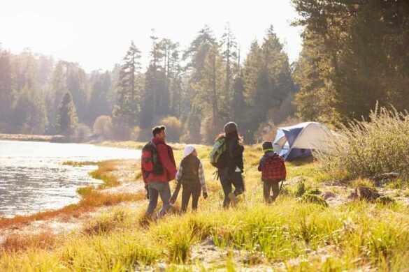 Une famille avec enfants et sacs à dos marche au bord d'une rivière vers une tente de camping, pour un trip mémorable en nature.