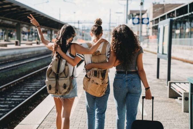 Trois jeunes femmes avec sacs à dos marchent bras dessus bras dessous sur un quai de gare, le début d'un trip mémorable.