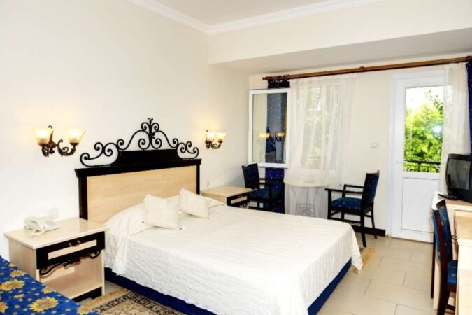 Chambre d'hôtel classique avec grand lit blanc, tête de lit en fer forgé et balcon donnant sur la verdure, typique des hôtels en ligne accessibles à petit budget.