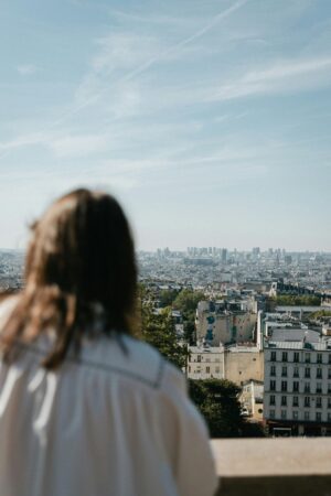 Une femme de dos contemple une vue panoramique de Paris sous un ciel clair. L'image capture le sentiment de revenir dans une destination déjà visitée.